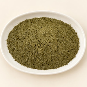 May include: A mound of finely ground, dark olive-green herbal powder centered on a smooth, white, oval-shaped ceramic dish. The texture of the powder appears uniform and dry, filling the basin of the plate while maintaining a rounded, conical shape.