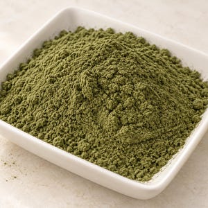 May include: A square white ceramic bowl filled with a mound of fine, olive-green powder. The powder has a dry, textured appearance with small clumps throughout, contrasting against the smooth, glossy surface of the white dish.