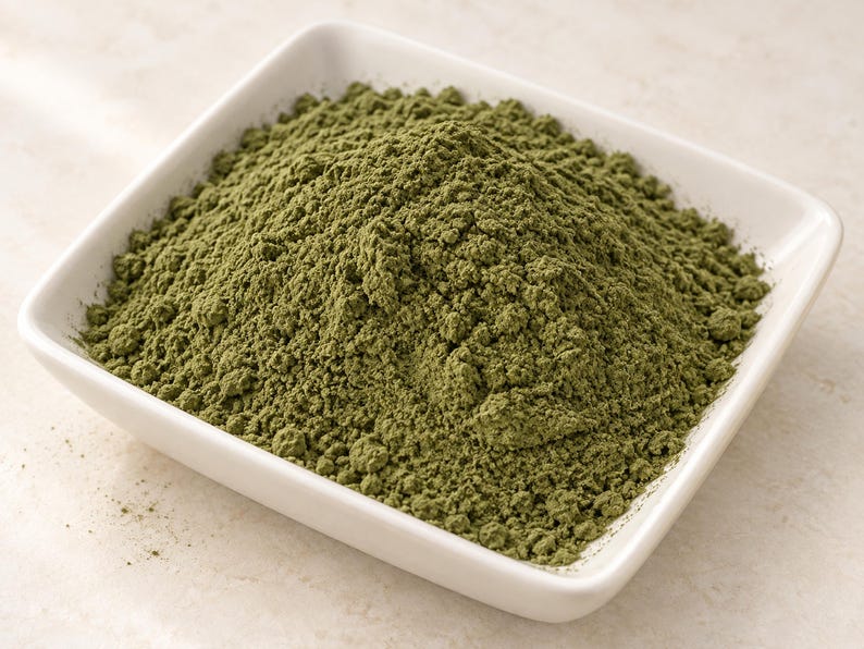 May include: A square white ceramic bowl filled with a mound of fine, olive-green powder. The powder has a dry, textured appearance with small clumps throughout, contrasting against the smooth, glossy surface of the white dish.
