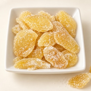 May include: A square white bowl filled with golden yellow slices of crystallized ginger coated in granulated sugar. Two additional pieces of the sugar-dusted ginger are positioned outside the bowl on the surface below the container.