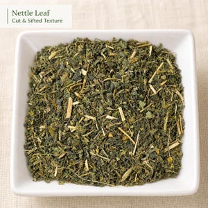 May include: A square white bowl filled with dried, cut, and sifted green nettle leaves. The text Nettle Leaf Cut & Sifted Texture is visible in the upper left corner. The dried herb mixture consists of small dark green leaf fragments and light tan stems.