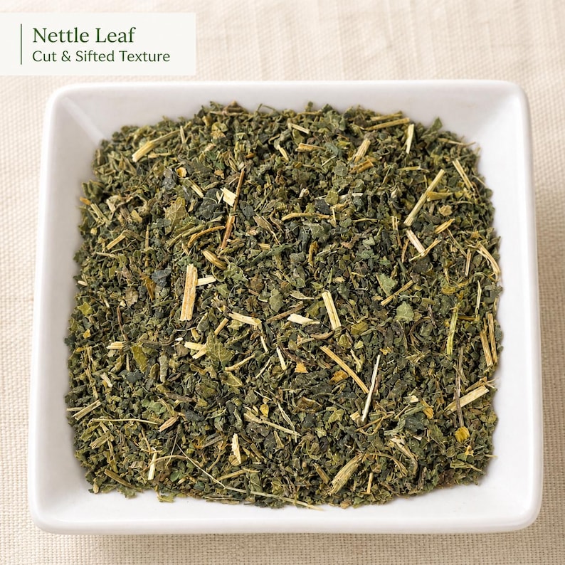 May include: A square white bowl filled with dried, cut, and sifted green nettle leaves. The text Nettle Leaf Cut & Sifted Texture is visible in the upper left corner. The dried herb mixture consists of small dark green leaf fragments and light tan stems.