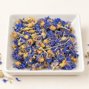 Op de afbeelding: Een vierkante witte keramische kom gevuld met een mengsel van gedroogde blauwe korenbloembladeren en lichtbruine bloemknoppen. Enkele losse blauwe bloemblaadjes en bloemhoofdjes liggen verspreid rond de basis van de kom.