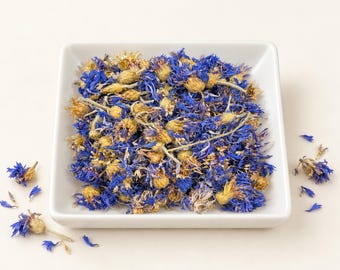 May include: A square white ceramic bowl filled with a mixture of dried blue cornflower petals and light tan flower buds. Several loose blue petals and flower heads are scattered around the base of the bowl on a neutral surface.