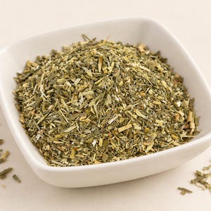 May include: A square white ceramic bowl filled with a mound of dried, finely chopped green herbs. The mixture consists of small, thin, fibrous pieces in varying shades of olive, sage, and light tan, with a few loose particles scattered nearby.