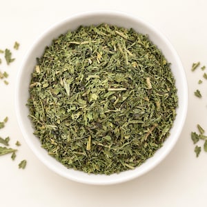 May include: A white ceramic bowl filled with dried green herbs, featuring a mix of finely chopped leaves and small stems. The texture is coarse and varied, with shades of dark and light green throughout the mixture, presented in a simple container.