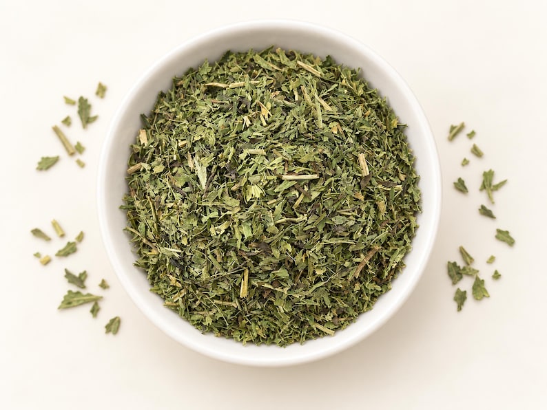 May include: A white ceramic bowl filled with dried green herbs, featuring a mix of finely chopped leaves and small stems. The texture is coarse and varied, with shades of dark and light green throughout the mixture, presented in a simple container.