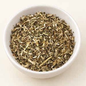 May include: A white ceramic bowl filled with a mixture of dried, chopped botanical herbs. The contents consist of small, fragmented pieces of green leaves, brown stems, and light tan fibrous plant matter, creating a textured, earthy appearance.