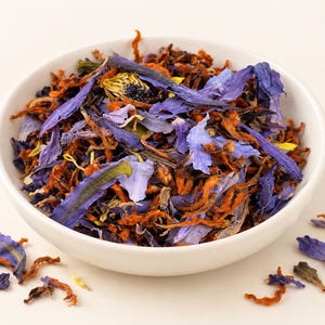 May include: A white ceramic bowl filled with a loose leaf herbal tea blend consisting of dried blue lotus flower petals, vibrant orange botanical strands, and small yellow floral pieces, with a few loose petals scattered around the base.