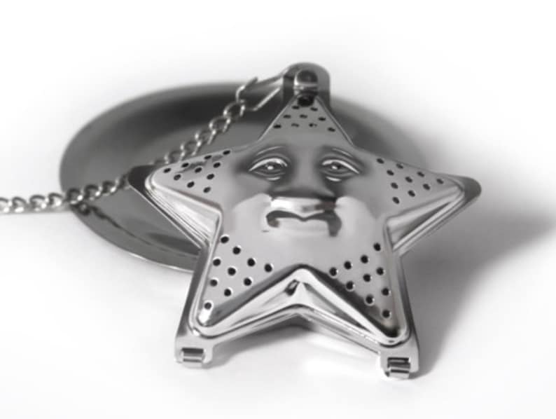 May include: A silver-colored metal tea infuser shaped like a five-pointed star. The front surface features a molded face with eyes, a nose, and a mouth, and is perforated with small holes for steeping tea. A metal chain is attached to the top.
