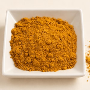 May include: A square white ceramic bowl filled with a mound of finely ground, vibrant golden-yellow turmeric powder. The spice has a dry, powdery texture with small clumps, and a small amount of the loose powder is scattered outside the bowl.