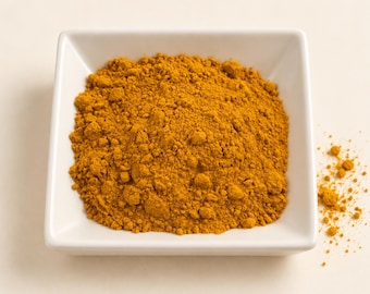 May include: A square white ceramic bowl filled with a mound of finely ground, vibrant golden-yellow turmeric powder. The spice has a dry, powdery texture with small clumps, and a small amount of the loose powder is scattered outside the bowl.