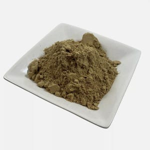 May include: A mound of fine, light brown, earthy-toned powder piled in the centre of a square, white ceramic dish. The powder has a slightly granular texture with small, loose clumps visible throughout the heap, resting on the smooth surface.