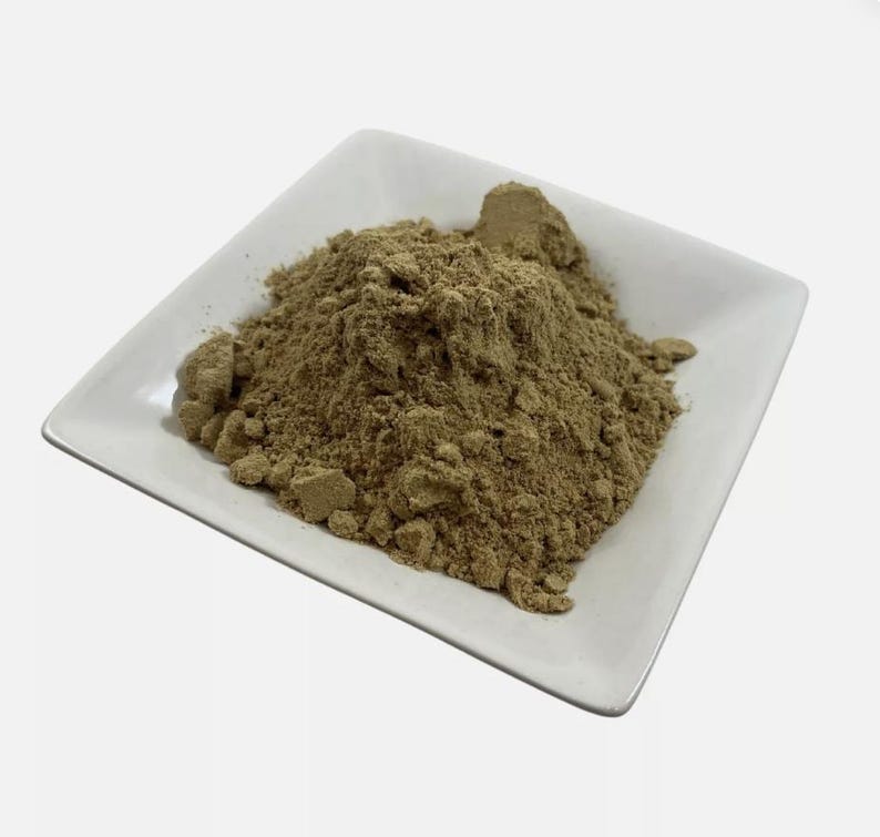 May include: A mound of fine, light brown, earthy-toned powder piled in the centre of a square, white ceramic dish. The powder has a slightly granular texture with small, loose clumps visible throughout the heap, resting on the smooth surface.