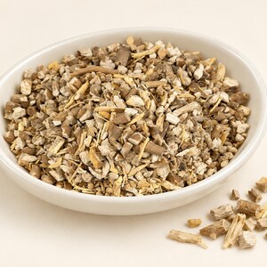 May include: A white ceramic bowl filled with a mixture of dried, chopped botanical root pieces in shades of light tan, beige, and dark brown. A small pile of the same dried root fragments is scattered outside the bowl on the surface.