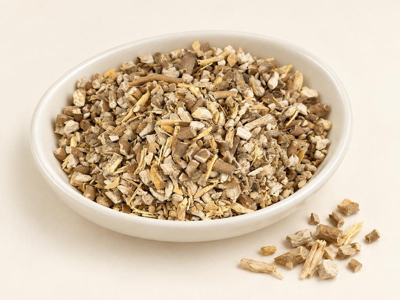 May include: A white ceramic bowl filled with a mixture of dried, chopped botanical root pieces in shades of light tan, beige, and dark brown. A small pile of the same dried root fragments is scattered outside the bowl on the surface.