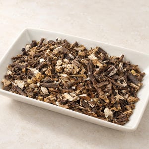 May include: A rectangular white ceramic dish filled with a heap of dried, chopped dark brown and tan root pieces. The mixture consists of irregular, woody fragments with varying textures, ranging from deep brown bark-like bits to lighter beige interiors.