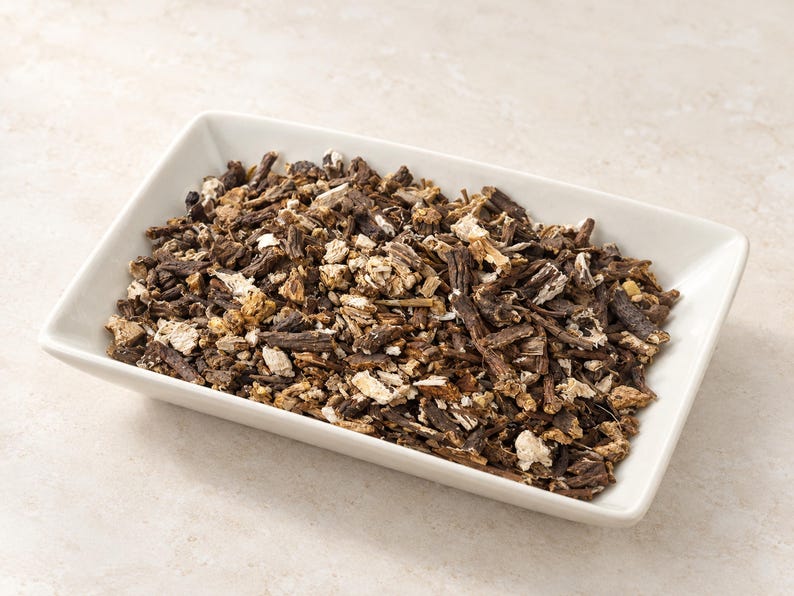 May include: A rectangular white ceramic dish filled with a heap of dried, chopped dark brown and tan root pieces. The mixture consists of irregular, woody fragments with varying textures, ranging from deep brown bark-like bits to lighter beige interiors.