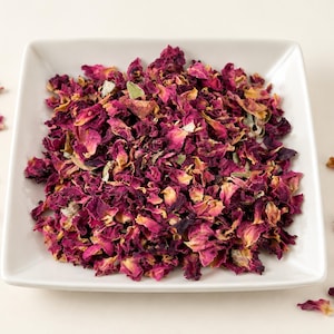 May include: A square white ceramic dish filled with a mound of dried rose petals in shades of deep magenta, dark pink, and light yellow. Several individual dried rose petals are scattered around the base of the dish on a plain surface.