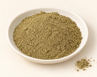 Puede incluir: Un montón de polvo herbal fino de color verde oliva contenido en un cuenco de cerámica blanco, redondo y poco profundo. Un pequeño montón separado del mismo polvo de textura terrosa se encuentra justo fuera del borde del cuenco.