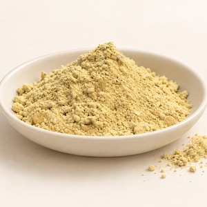 May include: A mound of fine, pale yellow powder piled in a shallow, round, off-white ceramic bowl. The texture of the powder appears dry and slightly granular, with a small amount of the substance scattered outside the bowl on the surface.