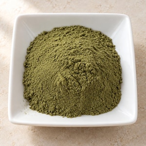 May include: A mound of fine, olive-green powder contained within a square, white ceramic bowl. The powder has a dry, matte texture and is piled high in the center of the dish, filling the basin while maintaining a distinct, granular appearance.