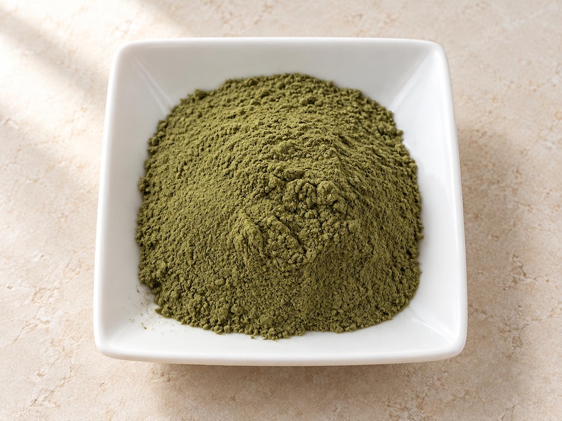 May include: A mound of fine, olive-green powder contained within a square, white ceramic bowl. The powder has a dry, matte texture and is piled high in the center of the dish, filling the basin while maintaining a distinct, granular appearance.