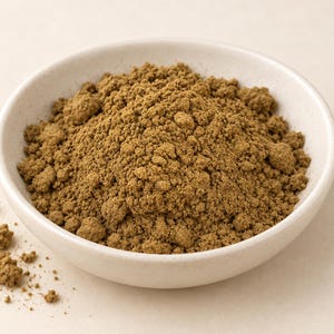 Pode incluir: Uma pequena tigela de cerâmica redonda, de cor esbranquiçada, cheia com um monte de especiarias em pó castanho-terra finamente moído. A textura do pó é granular e irregular, com alguns pequenos grumos espalhados pela tigela.