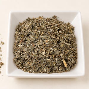 May include: A square white ceramic bowl filled with a mound of dried, crushed green and brown herbal leaves. The texture of the mixture is coarse and fragmented, featuring varied shades of olive, sage, and earthy brown tones throughout the pile.