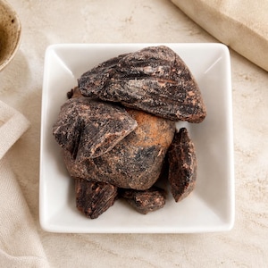 May include: A collection of dark brown, irregularly shaped chunks of raw resin or incense pieces piled inside a square white ceramic bowl. The pieces have a rough, textured surface with visible variations in color, ranging from deep black to reddish-brown.