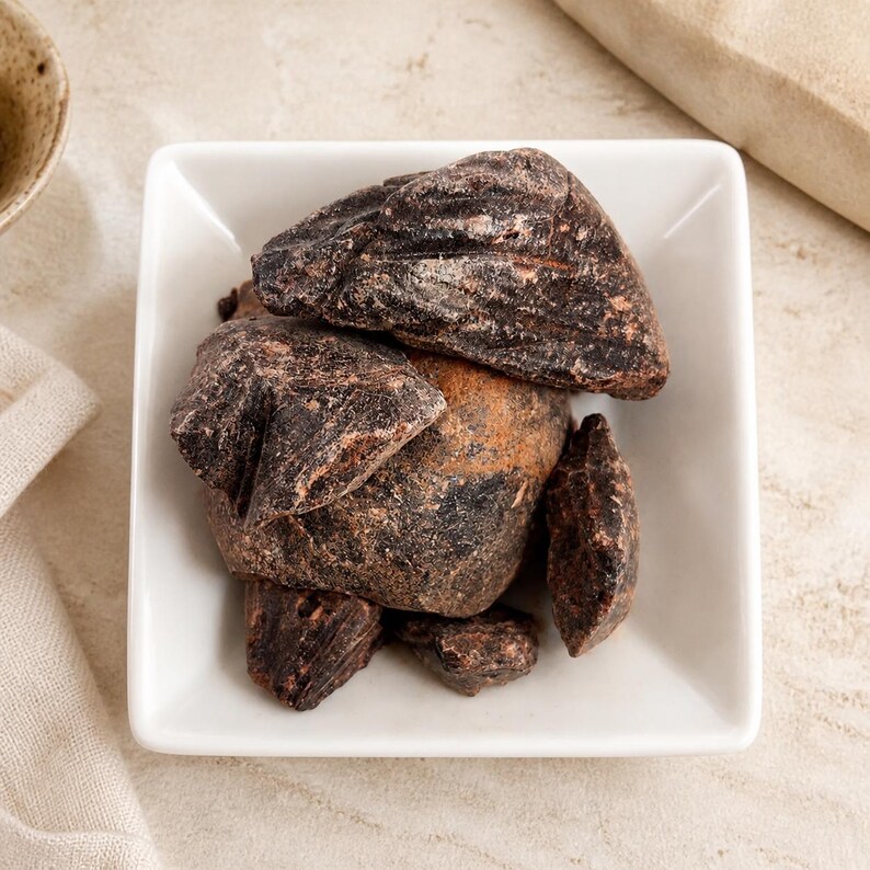 May include: A collection of dark brown, irregularly shaped chunks of raw resin or incense pieces piled inside a square white ceramic bowl. The pieces have a rough, textured surface with visible variations in color, ranging from deep black to reddish-brown.
