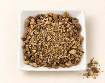 May include: A square white bowl filled with a mixture of dried, chopped brown root pieces and fine herbal particles. The texture is coarse and woody, featuring varying shades of tan, light brown, and dark brown fragments of organic plant matter.