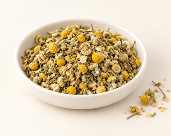 May include: A white ceramic bowl filled with dried chamomile flowers. The mixture consists of small, round, bright yellow flower heads and pale, off-white dried petals with thin green stems. A few loose chamomile flowers are scattered nearby.