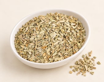 May include: A white ceramic bowl filled with dried, chopped green and tan herbal leaves. The mixture consists of small, irregular pieces of plant matter with varying shades of sage green, olive, and light brown, with a small pile of herbs nearby.