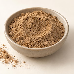 Può includere: Una ciotola in ceramica beige chiaro riempita con un mucchio di polvere fine color marrone chiaro. La consistenza della polvere appare asciutta e granulosa, con una piccola quantità di sostanza versata fuori dalla ciotola in basso a sinistra.