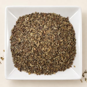 May include: A square white ceramic bowl filled with a mound of dried, crushed green herbs. The texture of the herbs is coarse and varied, featuring shades of dark olive, brown, and light green. A few small herb particles are scattered outside the bowl.