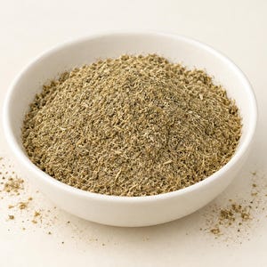 May include: A white ceramic bowl filled with a mound of finely ground, light brown herbal powder. The texture of the powder appears fibrous and dry, with small particles visible throughout the mixture. A few loose grains are scattered outside the bowl.