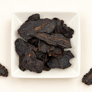May include: A square white ceramic bowl filled with several dark brown, dried, irregular-shaped herbal root slices. The slices have a rough, textured surface with visible fibrous brown streaks running through the dark, matte material.