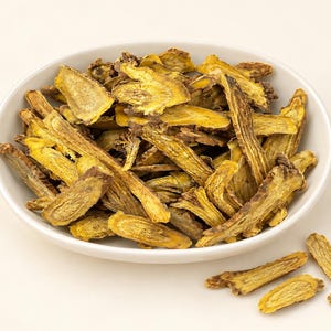 May include: A white ceramic bowl filled with numerous dried, sliced pieces of yellow and brown root. The slices have a fibrous, woody texture with a bright yellow interior and a darker, rougher outer bark, with a few loose pieces resting nearby.