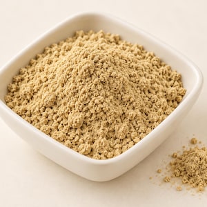 May include: A square white ceramic bowl filled with a mound of fine, light tan, powdery substance. The texture of the powder appears granular and dry, with a small separate pile of the same light tan material resting outside the bowl.