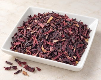 May include: A square white ceramic bowl filled with a mound of dried hibiscus flower petals. The petals are deep burgundy and dark red in color with some lighter tan-colored fibrous pieces mixed throughout the pile of loose herbal tea leaves.