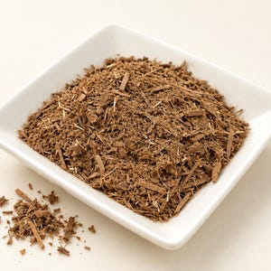 May include: A white square ceramic bowl filled with a pile of finely shredded brown bark or dried root material. The texture is coarse and fibrous, consisting of small, thin, splinter-like pieces in various shades of light and dark brown.