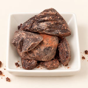 May include: A square white ceramic bowl filled with several irregular, dark brown, solid chunks of raw cacao. The pieces have a rough, matte texture with visible variations in color, ranging from deep espresso to lighter reddish-brown tones.