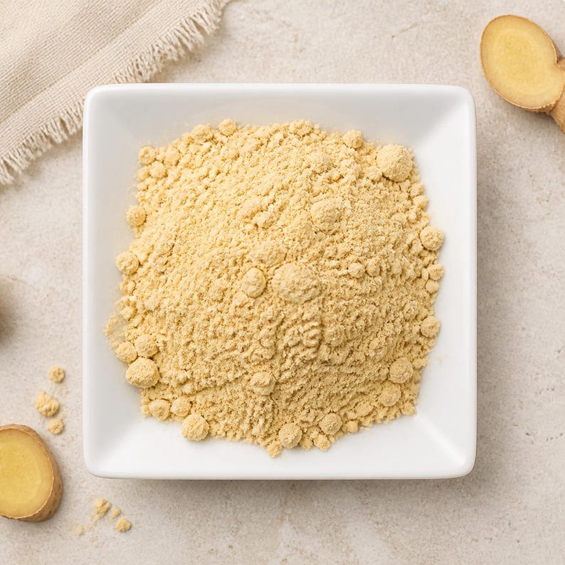 May include: A square white bowl filled with a mound of fine, pale yellow ginger powder. The texture of the powder is slightly clumpy and granular. Two sliced pieces of fresh, light brown ginger root are positioned near the corners of the bowl.