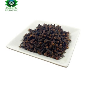 May include: A white square plate filled with dried whole cloves. The cloves are dark brown and have a slightly reddish hue.