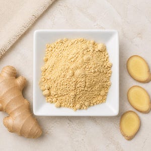 May include: A square white bowl filled with light tan ginger powder, positioned next to a whole ginger root and three thin, circular slices of fresh ginger root. The texture of the powder is fine and granular, contrasting with the smooth bowl.
