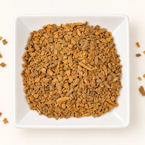 May include: A square white bowl filled with a mound of small, irregular, golden-brown cinnamon bark pieces. The texture of the spice is coarse and woody, with varying shades of tan and brown throughout the pile, and a few scattered fragments.