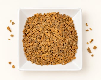May include: A square white bowl filled with a mound of small, irregular, golden-brown cinnamon bark pieces. The texture of the spice is coarse and woody, with varying shades of tan and brown throughout the pile, and a few scattered fragments.