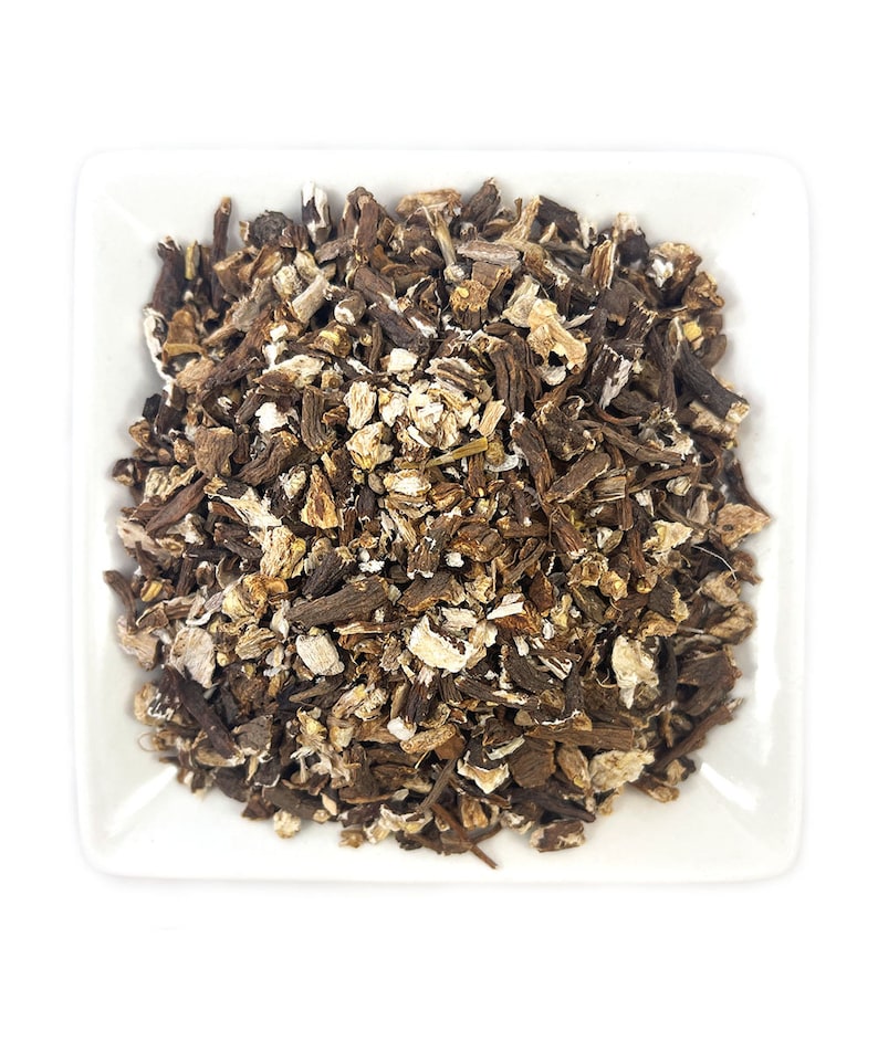 May include: A square white bowl filled with a mound of dried, chopped botanical root pieces. The mixture consists of small, irregular fragments in varying shades of dark brown, tan, and off-white, showcasing a rough, fibrous texture throughout.