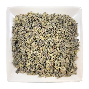 May include: A white square dish filled with loose green tea leaves. The tea leaves are a mix of whole leaves and broken pieces.